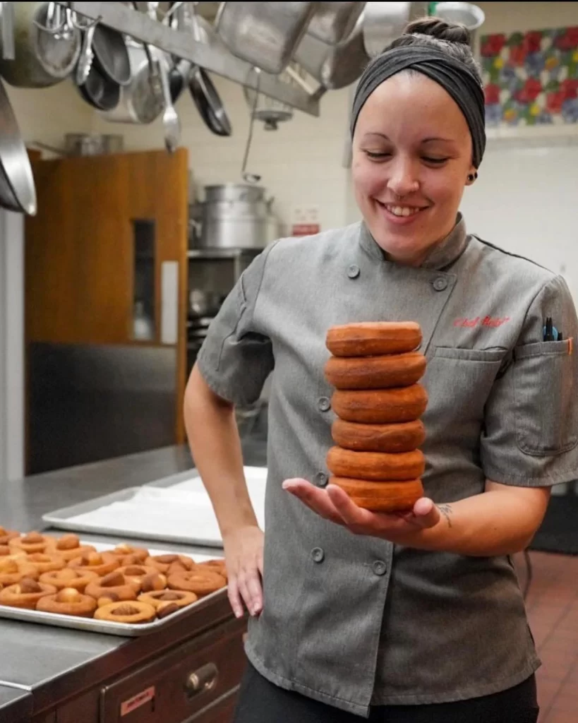 A chef in a kitchen smiles while balancing a stack of seven donuts on one hand, with more donuts in the background.